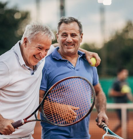 Two cheerful senior men talking while walking on the outdoor tennis court